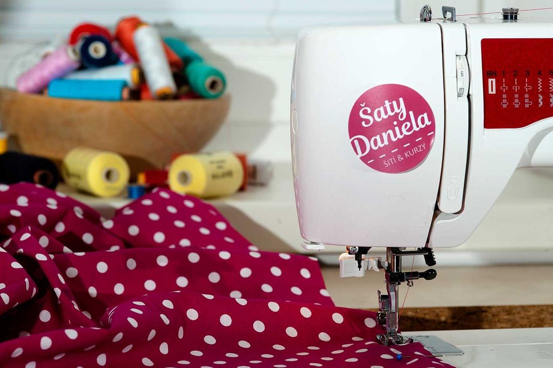 Šicí stroj se samolepkou Šaty Daniela, vedle cívky nití a puntíkovaná látka